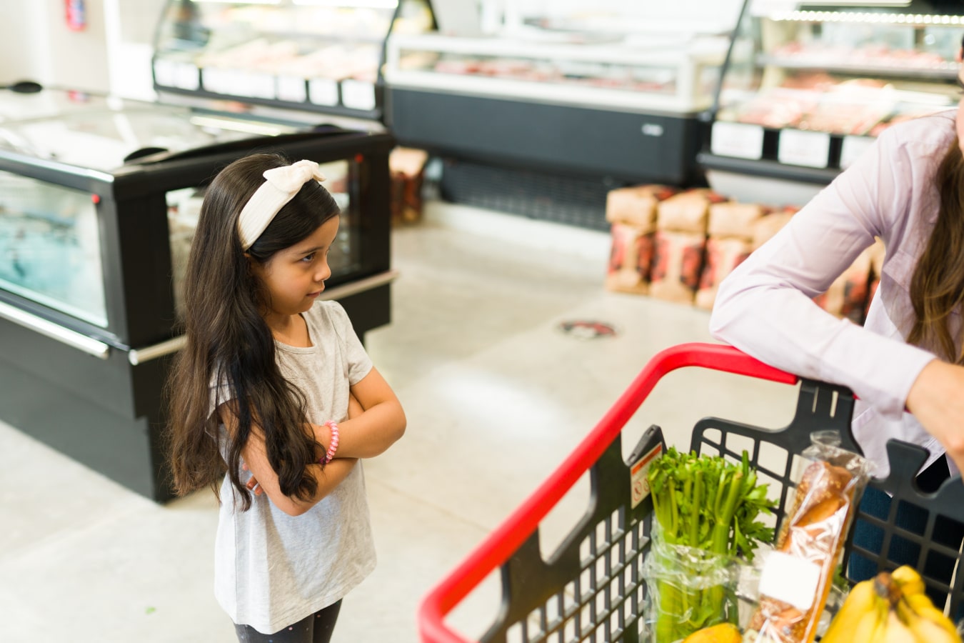 una niña enfadada está de pie junto a la cesta de la compra, está disgustada porque los padres no le quieren comprar algo